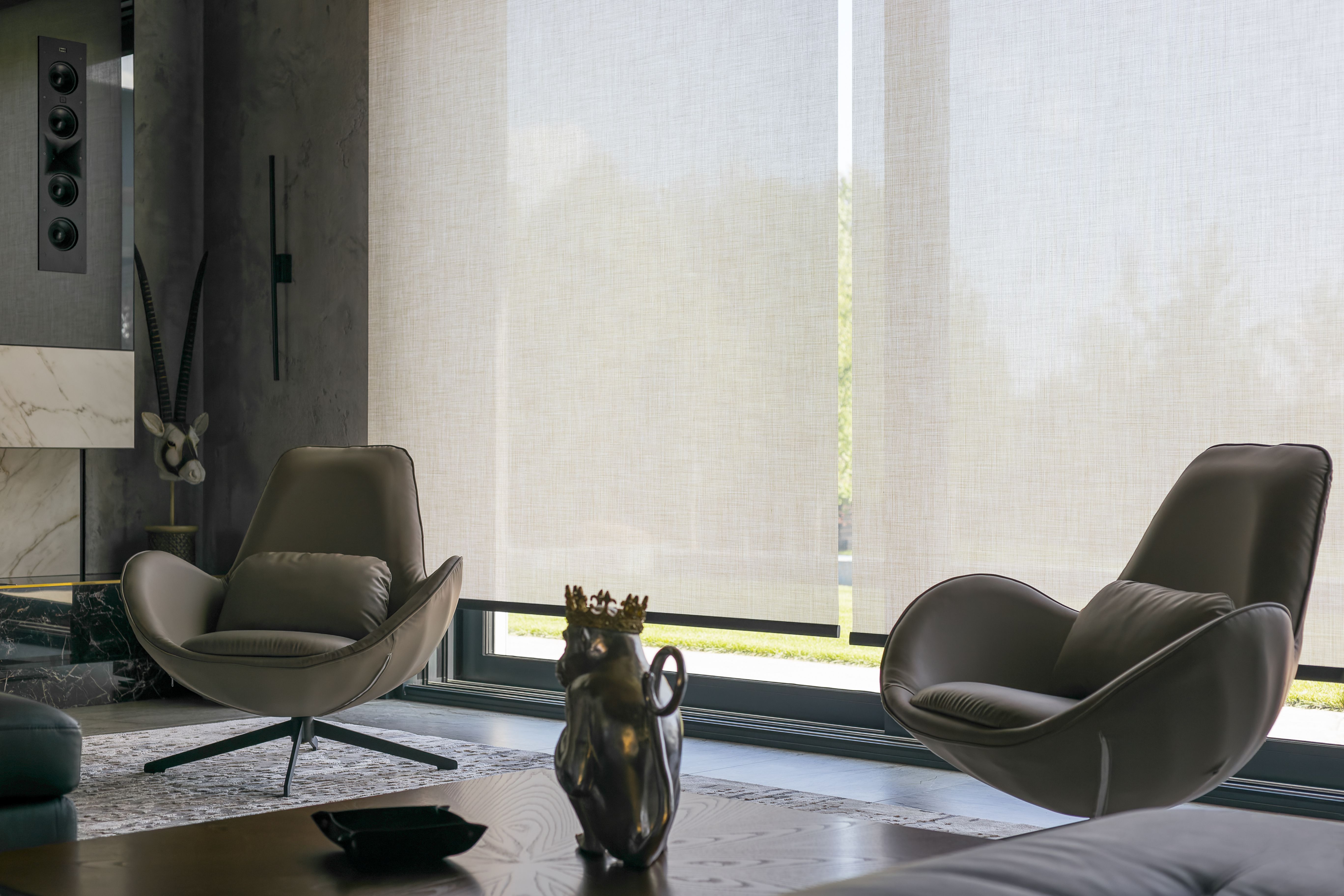 Modern living room with two contemporary lounge chairs facing a large window covered with light-filtering roller shades, a decorative sculpture of a crowned animal on a wooden coffee table, and wall-mounted speakers and art pieces enhancing the elegant, minimalist interior.