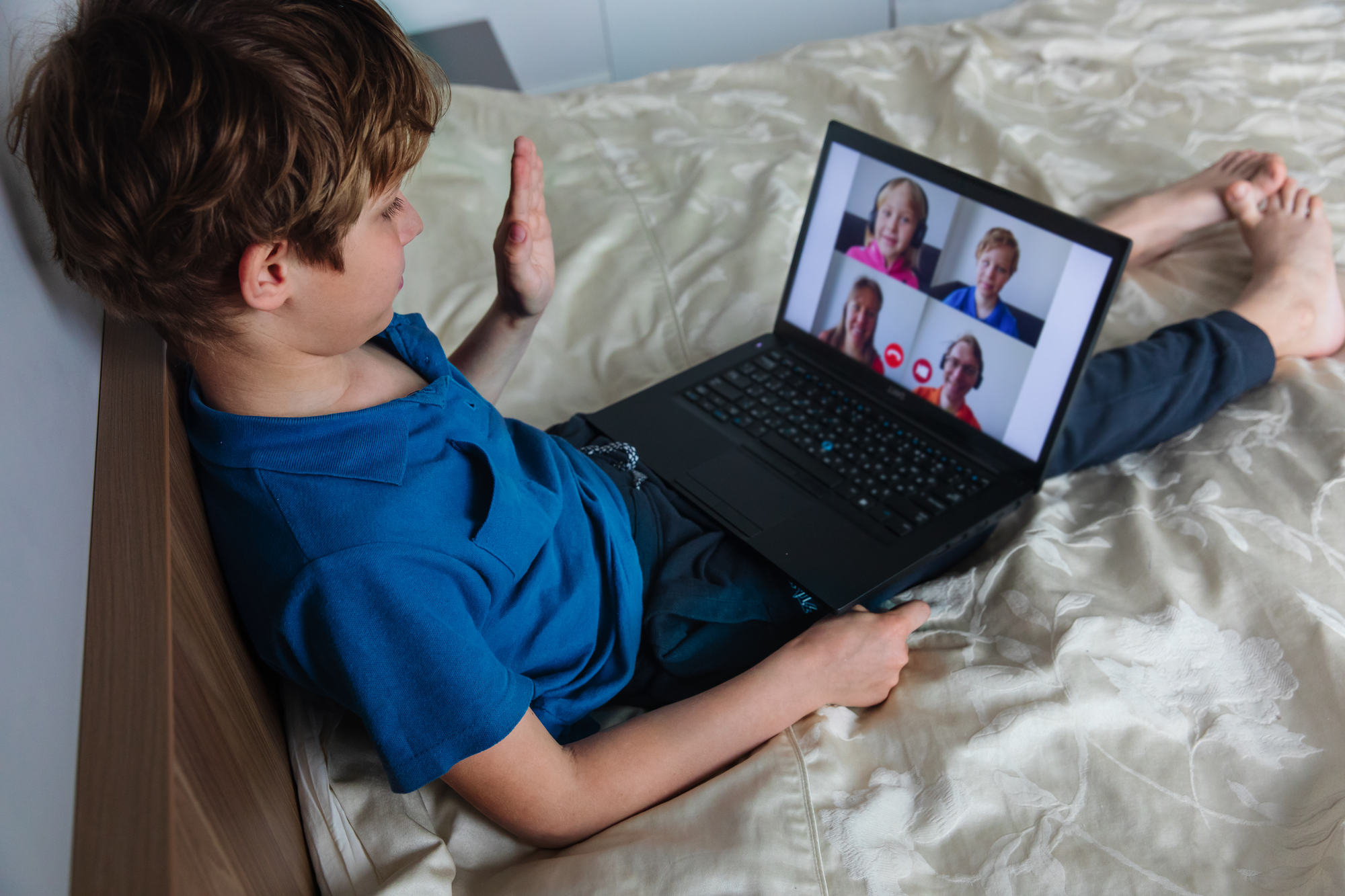Boy lying on a bed, waving during a group video call on a laptop with four people on screen.