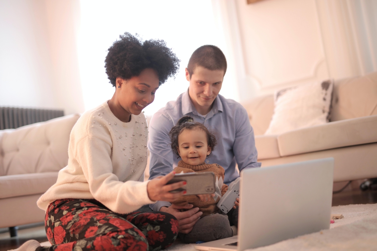 Smiling family of three sitting on the floor, using a laptop and taking a selfie together in a cozy living room.