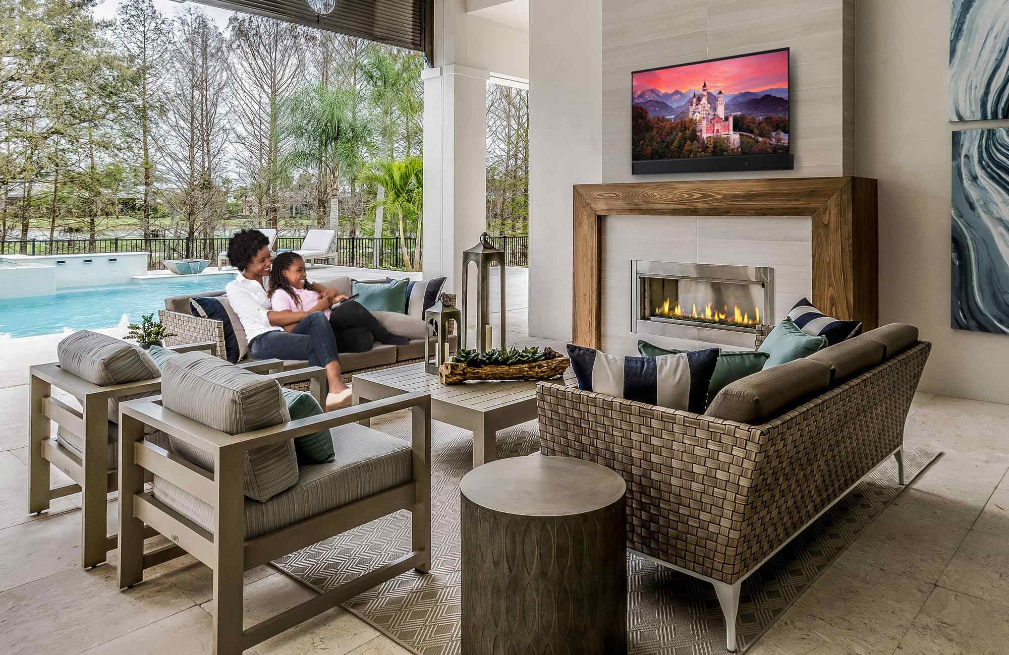 Mother and daughter watching an outdoor TV by the pool and fireplace on a covered patio