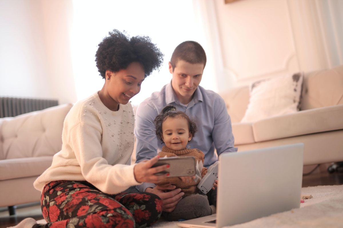 Smiling family of three sitting on the floor, using a laptop and taking a selfie together in a cozy living room.